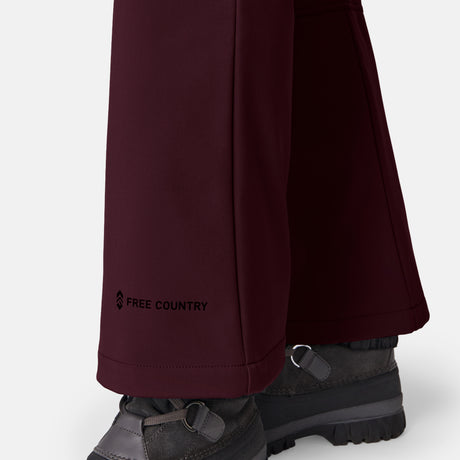 Close-up of a person wearing Free Country Women’s Stormlite Super Softshell® Ski Pant in dark burgundy with water resistance, four-way stretch, and logo on the leg. Gray hiking boots. Plain white background.