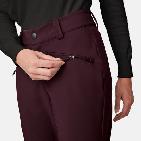 A person, visible from torso to hand, unzips a small front pocket on burgundy Women’s Stormlite Super Softshell® Ski Pants by Free Country, paired with a black long-sleeve shirt.