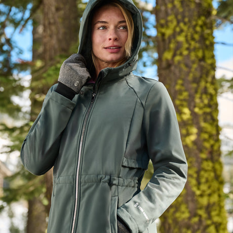 A woman wearing a sage green hooded outdoor jacket with gray knit gloves, standing outdoors in front of blurred trees.