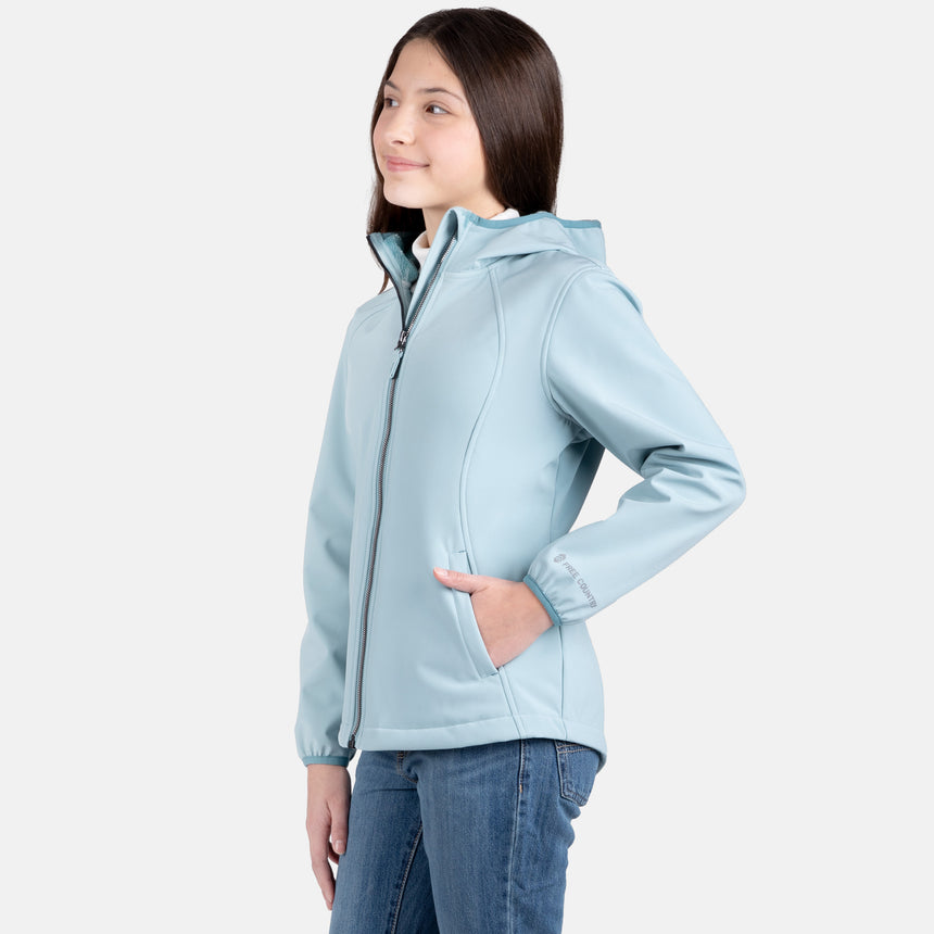 [Samantha is wearing size M.] Model wearing a Free Country light blue softshell zip-front hooded jacket with a smooth, weather-resistant finish, side pockets, and tonal stitching—layered over a white turtleneck and paired with blue jeans for a clean, casual winter look.