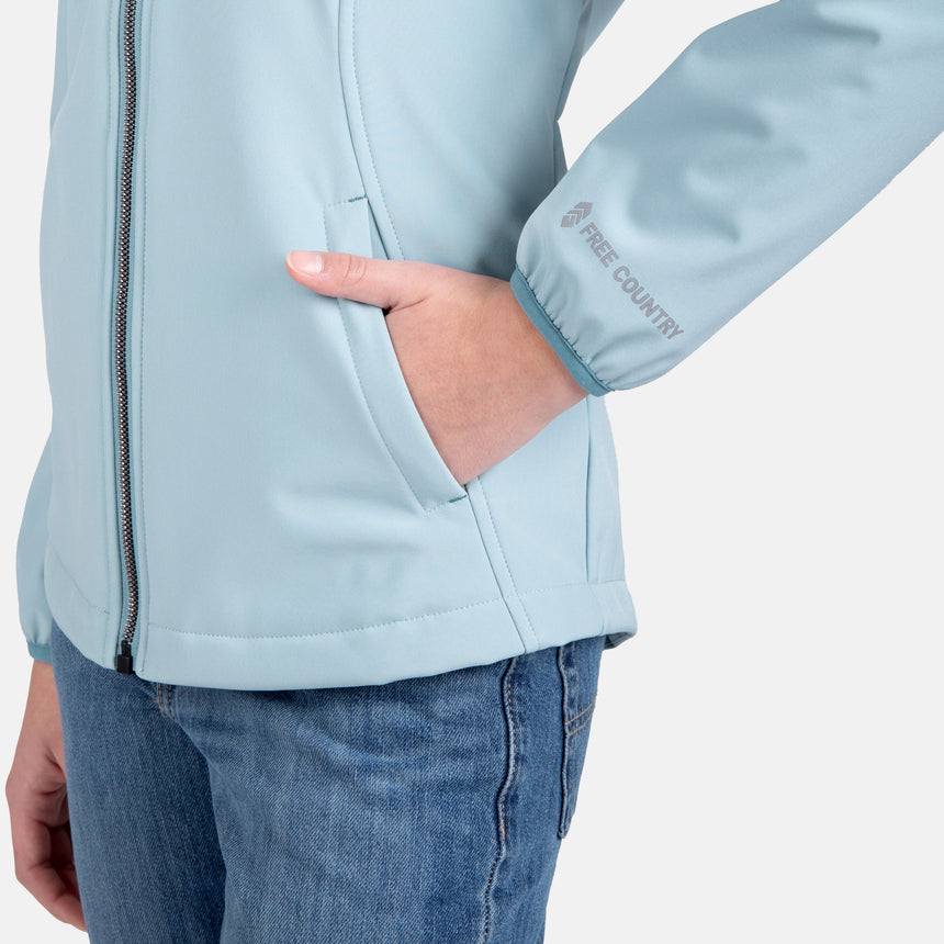 [Samantha is wearing size M.] Model wearing a Free Country light blue softshell zip-front hooded jacket with a smooth, weather-resistant finish, side pockets, and tonal stitching—layered over a white turtleneck and paired with blue jeans for a clean, casual winter look.
