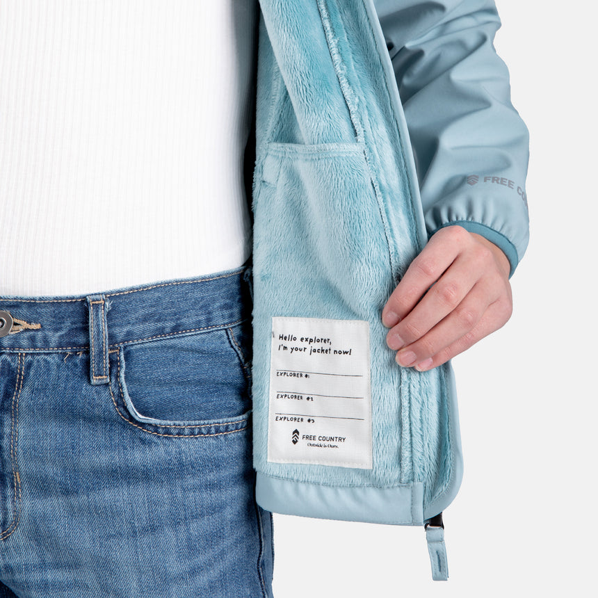[Samantha is wearing size M.] Model wearing a Free Country light blue softshell zip-front hooded jacket with a smooth, weather-resistant finish, side pockets, and tonal stitching—layered over a white turtleneck and paired with blue jeans for a clean, casual winter look.