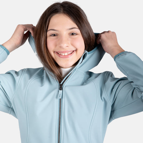 [Samantha is wearing size M.] Model wearing a Free Country light blue softshell zip-front hooded jacket with a smooth, weather-resistant finish, side pockets, and tonal stitching—layered over a white turtleneck and paired with blue jeans for a clean, casual winter look.