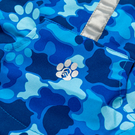 A close-up of Free Country’s Dog Raincoat shows blue camo waterproof fabric, a silver reflective strip, and a pocket decorated with a paw print featuring a small geometric logo.