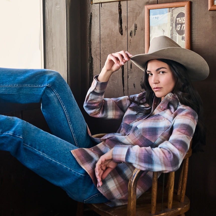 A woman reclining in a rustic wooden chair, wearing a brown cowboy hat, a purple and blue plaid flannel shirt, and blue jeans, set against a dark wood-paneled wall.