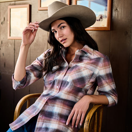 A woman wearing a brown cowboy hat and a long-sleeve plaid button-up shirt with pink, purple, and white tones, sitting on a wooden chair against a rustic wood-paneled wall with framed pictures.