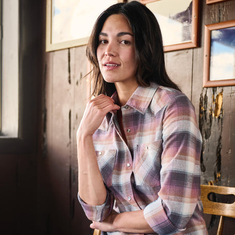 A woman wearing a long-sleeve plaid button-up shirt in muted pink, purple, and beige tones, posed in a rustic indoor setting with wooden walls and framed pictures.