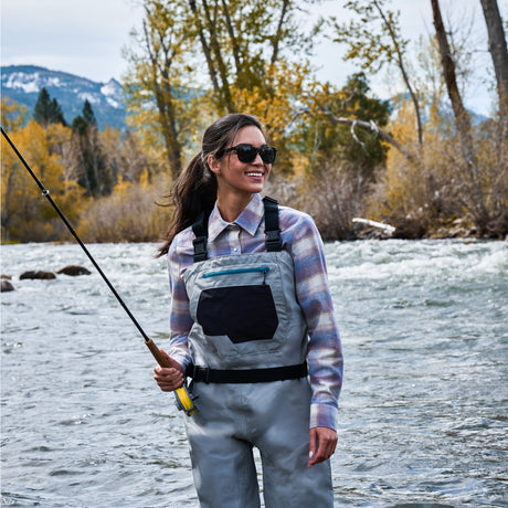 A woman wearing gray fly-fishing waders and a plaid shirt stands in a river holding a fishing rod, with mountains and autumn trees in the background.