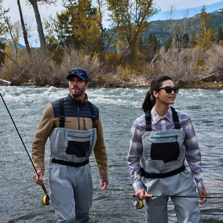 Two people wearing waders and holding fishing rods stand in a river with trees and mountains in the background. One of them is dressed warmly in the Free Country Men’s Frore Knit Fleece Jacket, which features a convertible collar—perfect for the cool, sunny day.