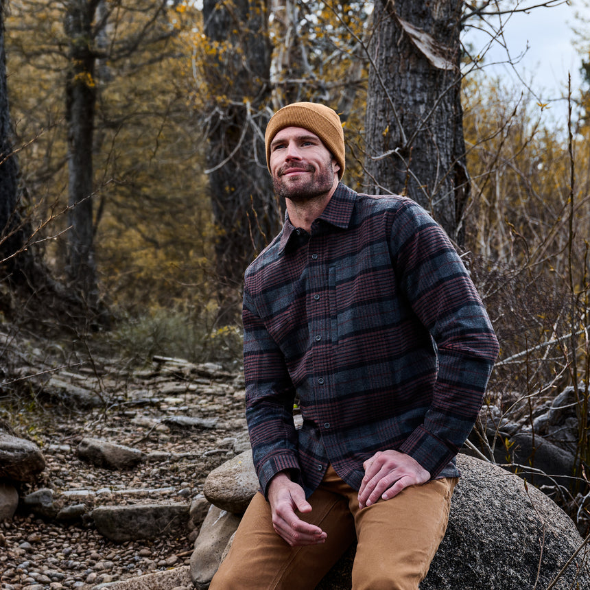 A man wearing a dark plaid button-up shirt and tan beanie sits on a large rock in a wooded outdoor setting.