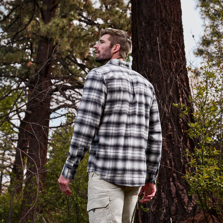 A man wearing a black and white plaid long-sleeve flannel shirt and khaki cargo pants, standing in a forest next to a large tree trunk.