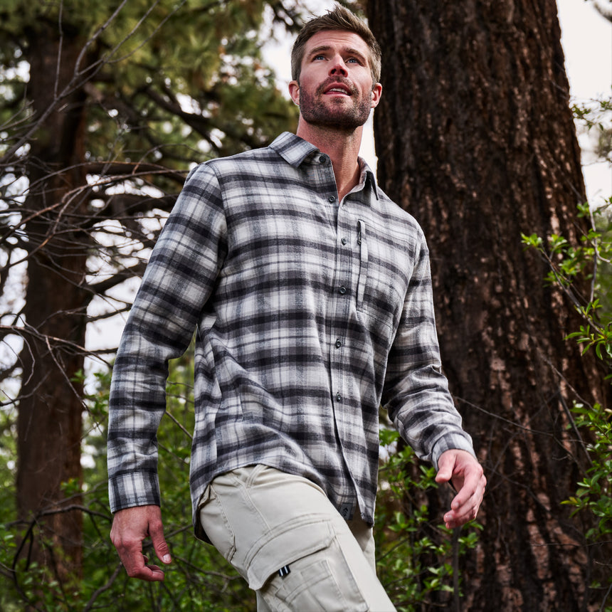 A man wearing a gray and black plaid long-sleeve button-up shirt and khaki cargo pants, standing in a forest with a large tree trunk behind him.