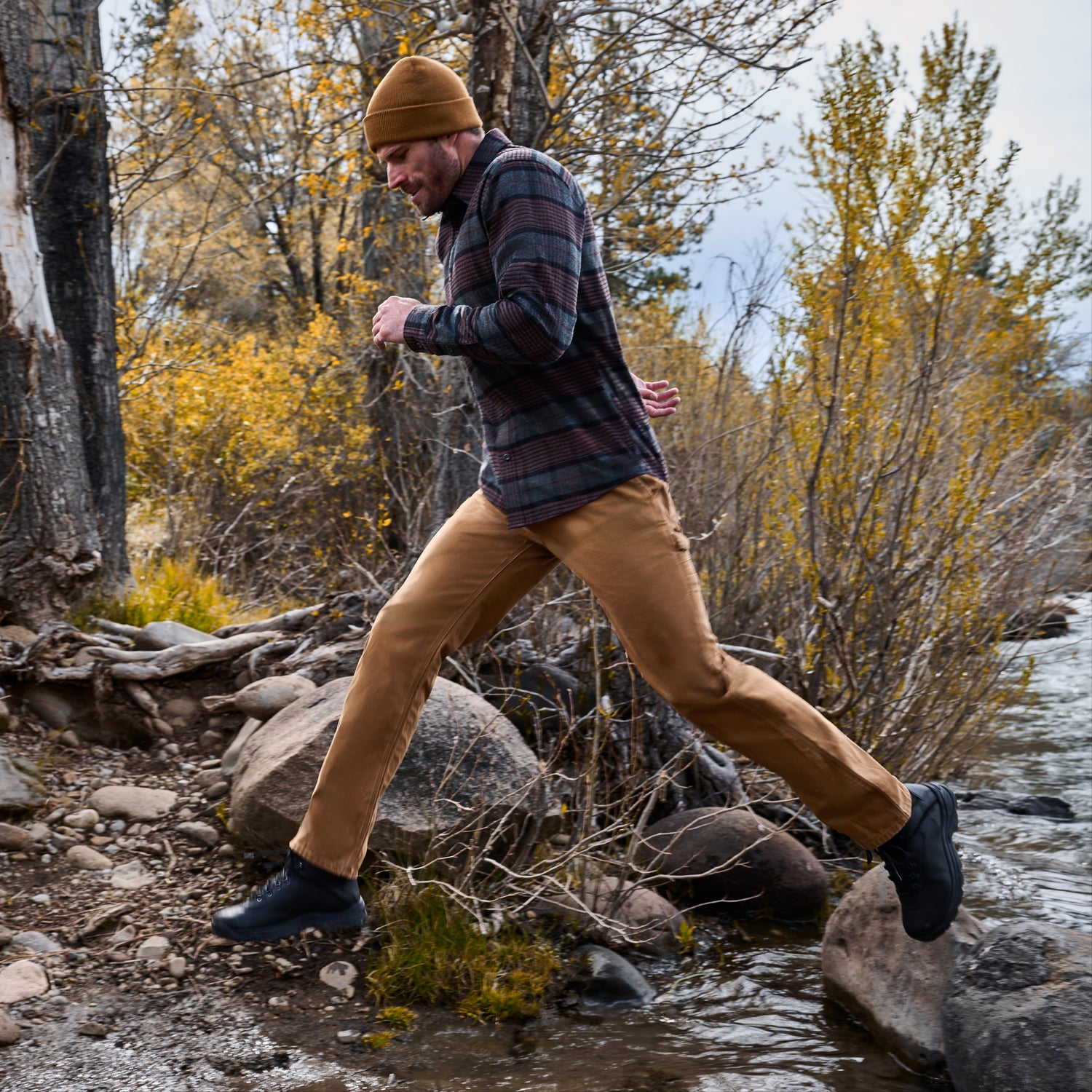 A man wearing a brown beanie, dark gray plaid shirt, tan pants, and black boots is running across rocks near a stream in an outdoor autumn setting with yellow foliage.