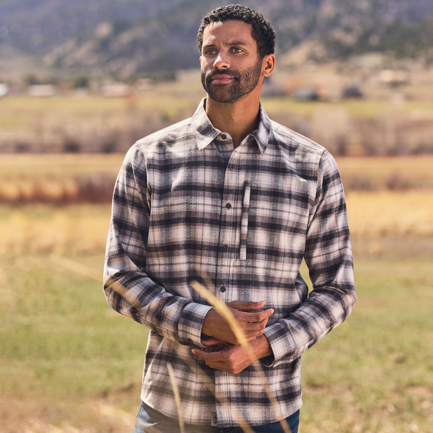 Man wearing a plaid shirt standing in a field with mountains in the background