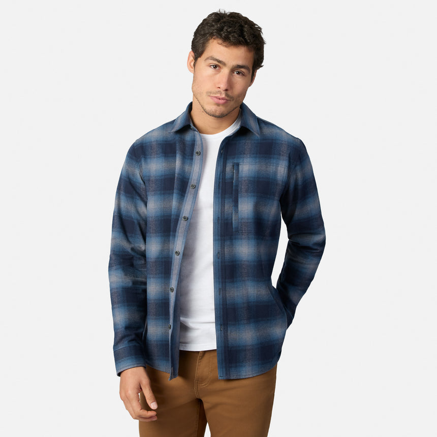 [Model is wearing size M.] A man wearing a blue and navy plaid button-down shirt over a white t-shirt and brown pants, standing against a light gray studio background.