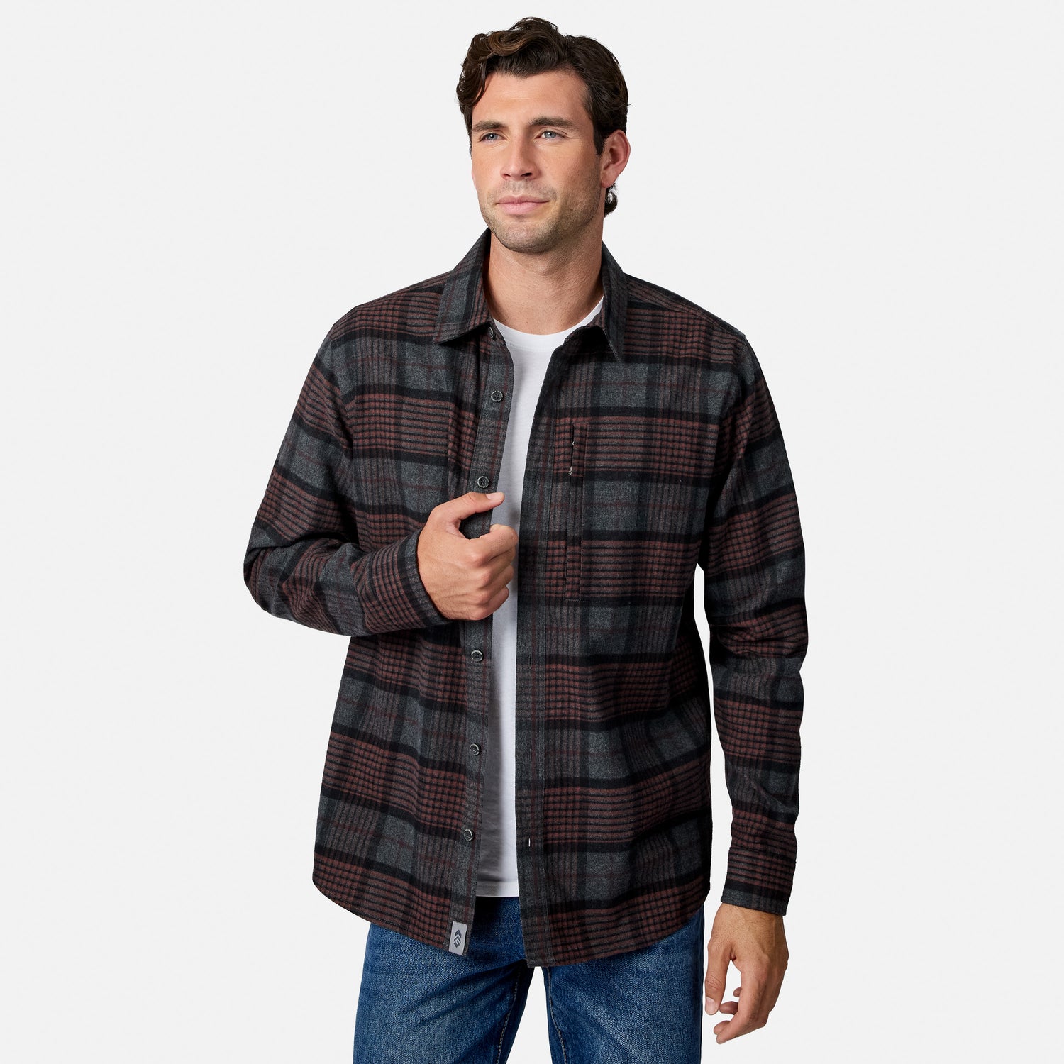 [Model is wearing size M.] A man wearing a dark gray and brown plaid button-down flannel shirt over a white t-shirt and blue jeans, standing against a light gray studio background.