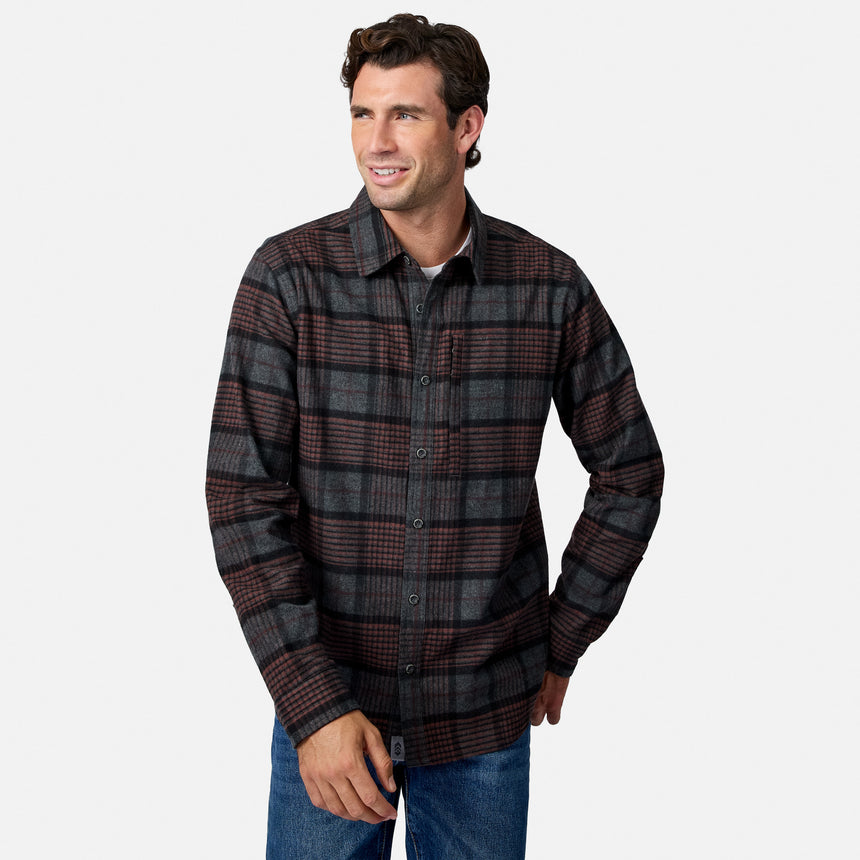 [Model is wearing size M.] A man wearing a dark brown and black plaid button-down flannel shirt with blue jeans, standing against a light gray background.