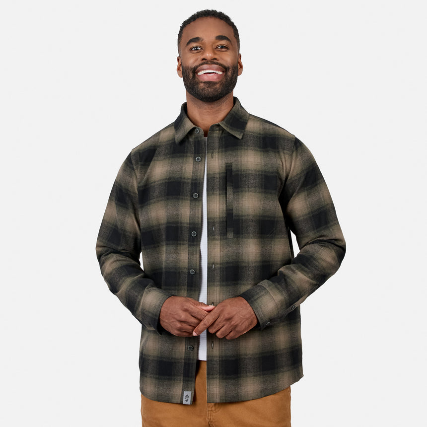 [Model is wearing size M.] A man wearing a long-sleeve button-up plaid overshirt in dark green and brown tones, layered over a white t-shirt and brown pants, against a light gray background.