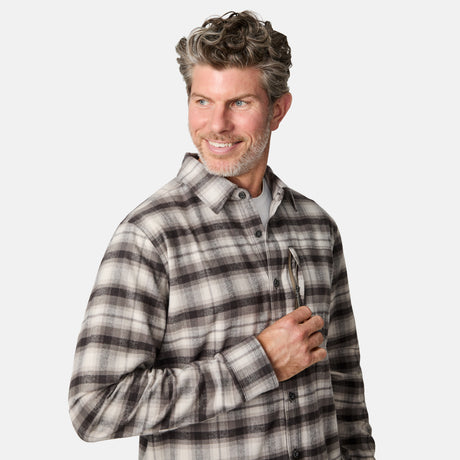 A smiling man with short, wavy gray hair and a beard wears the Free Country Men's Easywear Flannel Shirt in gray and white plaid, slipping sunglasses into his chest zipper pocket against a plain white background.