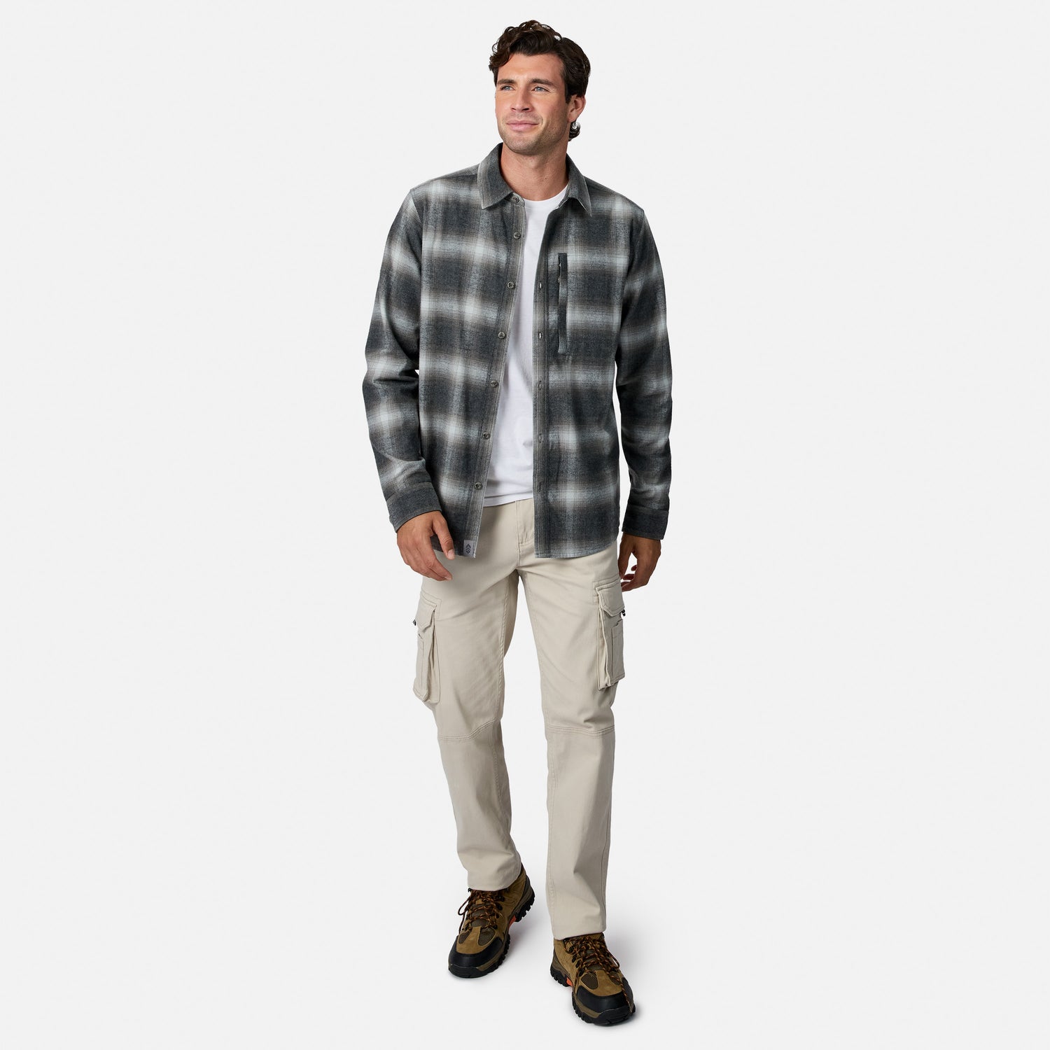 [Model is wearing size M.] A man modeling a dark gray and black plaid button-down flannel shirt over a white t-shirt, paired with beige cargo pants and brown hiking boots, on a light gray background.