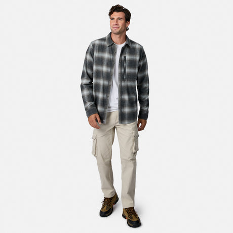 [Model is wearing size M.] A man modeling a dark gray and black plaid button-down flannel shirt over a white t-shirt, paired with beige cargo pants and brown hiking boots, on a light gray background.