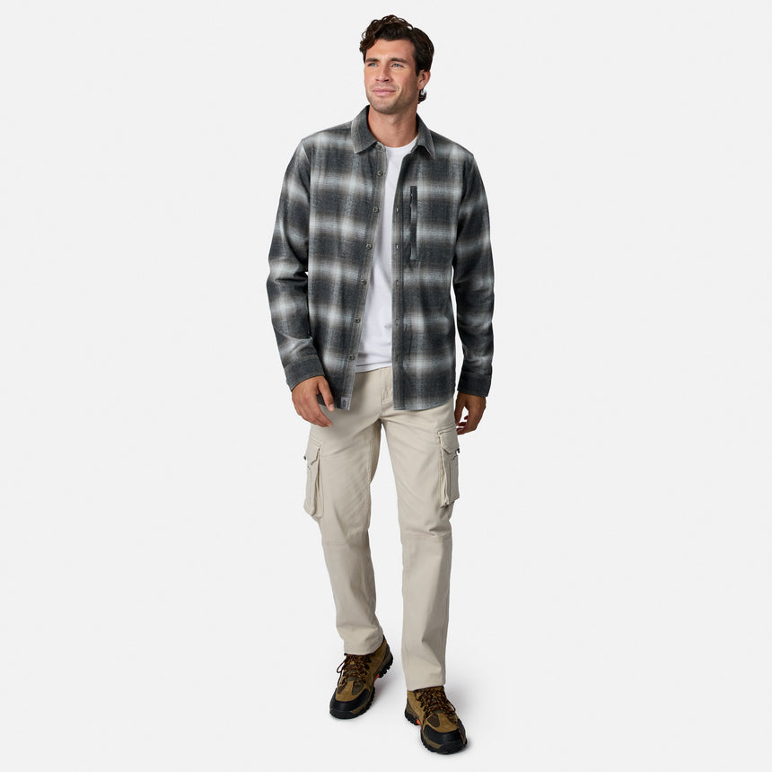 [Model is wearing size M.] A man modeling a dark gray and black plaid button-down flannel shirt over a white t-shirt, paired with beige cargo pants and brown hiking boots, on a light gray background.