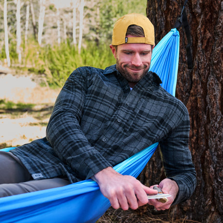 A man wearing a yellow cap and a dark gray plaid flannel shirt relaxes in a bright blue camping hammock strung between two trees in an outdoor forest setting.