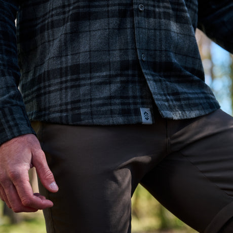Close-up of a person wearing a dark gray and black plaid flannel shirt with a small logo tab near the hem, paired with dark brown pants in an outdoor setting.