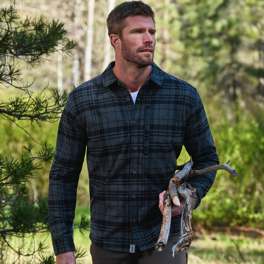 A man wearing a dark green and black plaid flannel shirt stands outdoors in a wooded setting, holding a piece of driftwood.