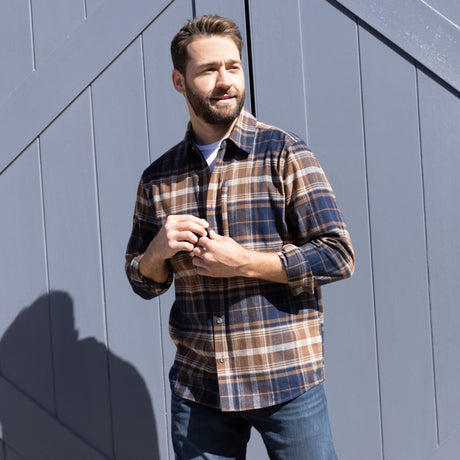 A bearded man stands outside in sunlight, smiling and clasping his hands, wearing the Free Country Men's Easywear II Fleece Lined Flannel Shirt with jeans, with a gray wooden fence in the background.
