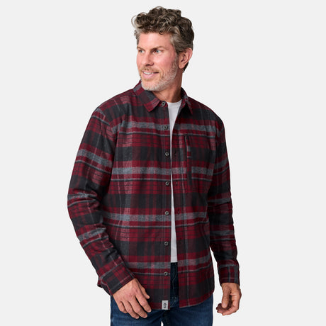 A man with wavy gray hair and a beard wears the Free Country Men's Easywear II Fleece Lined Flannel Shirt in red and black plaid over a white T-shirt and blue jeans, smiling as he stands against a plain light gray background.