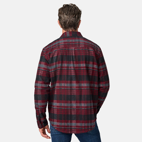 A man faces away from the camera wearing the Free Country Men's Easywear II Fleece Lined Flannel Shirt in a red, black, and gray plaid pattern with blue jeans, against a plain white background.