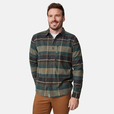A man with short brown hair and a beard is smiling while wearing the Free Country Men's Easywear II Fleece Lined Flannel Shirt over a white undershirt and brown pants, standing against a plain light background.