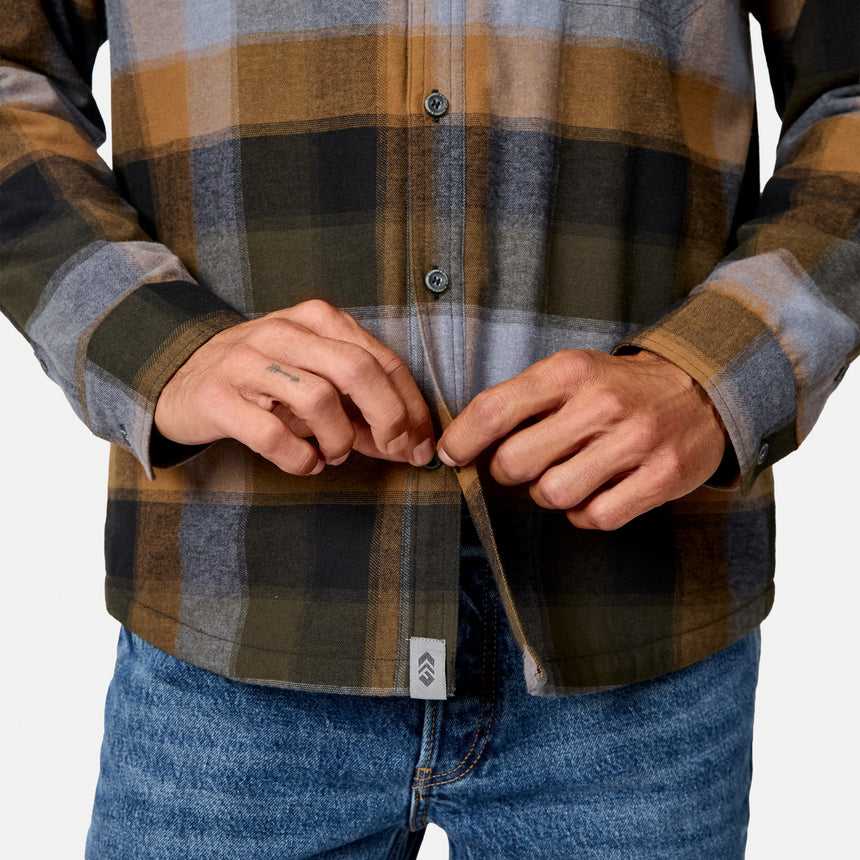 A close-up of a person wearing a long-sleeve plaid flannel shirt in brown, gray, and black, with blue jeans, adjusting the front placket.