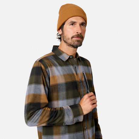 [Model is wearing size M.] A man wearing a mustard yellow beanie and a brown, gray, and green plaid flannel shirt, holding a pair of glasses against a light gray background.