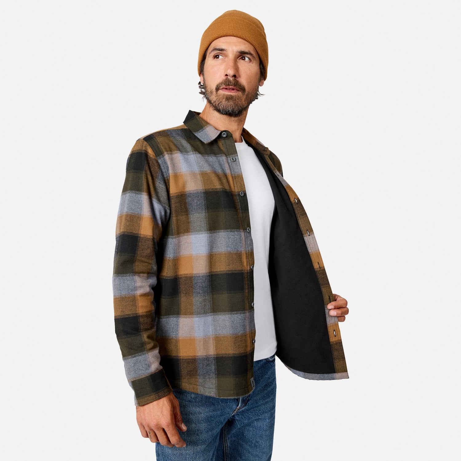 [Model is wearing size M.] A man wearing a brown, black, and gray plaid flannel shirt over a white t-shirt and blue jeans, with a tan beanie, holding the shirt open to show the black lining against a light gray background.