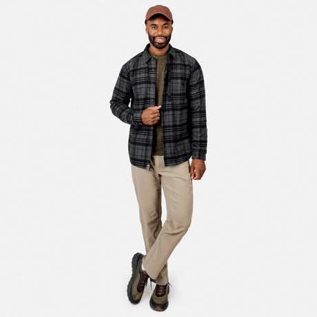 [Model is wearing size M.] A man modeling a dark gray and black plaid button-down shirt over an olive green t-shirt, paired with beige pants, hiking boots, and a brown baseball cap, against a light gray background.