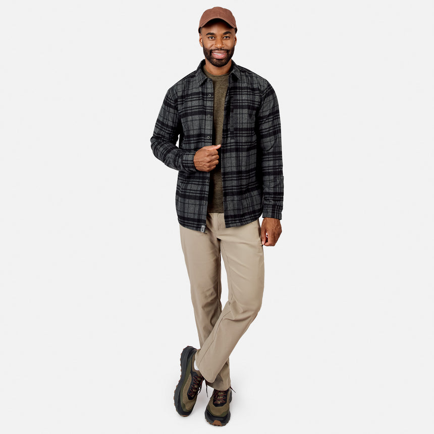 [Model is wearing size M.] A man modeling a dark gray and black plaid button-down shirt over an olive green t-shirt, paired with beige pants, hiking boots, and a brown baseball cap, against a light gray background.