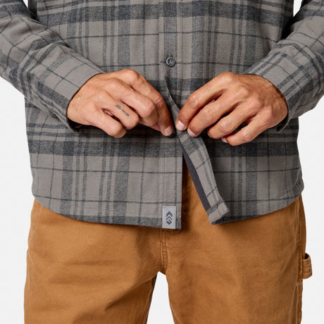 [Model is wearing size M.] A close-up of a person wearing a gray and black plaid button-up shirt, adjusting the front placket with both hands, paired with tan pants against a clean white background.