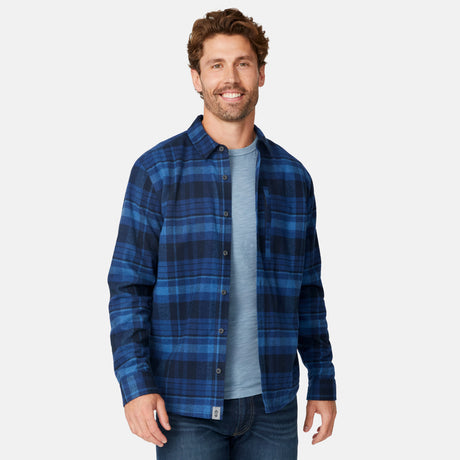 A man with brown hair and a beard smiles, wearing the Free Country Men's Easywear II Fleece Lined Flannel Shirt over a light blue t-shirt and jeans, standing against a plain white background.