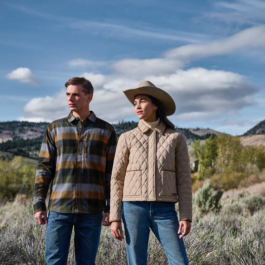 Man in a plaid shirt jacket and woman in a quilted jacket standing in open rangeland
