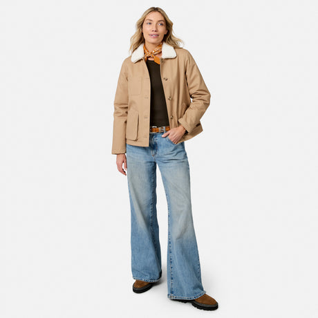 [Model is wearing size S.] A woman modeling a tan waxed canvas utility jacket with a white shearling-style collar, worn over a black top and blue wide-leg jeans, against a plain white studio background.