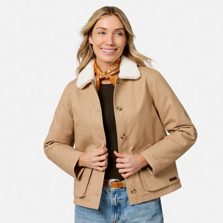 [Model is wearing size S.] A woman modeling a tan waxed canvas utility jacket with a white sherpa collar, styled with a patterned orange scarf, black top, and blue jeans against a light gray background.