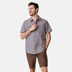 A man smiles with one hand in his pocket and the other adjusting his shirt, wearing the Men's Expedition Nylon Rip-Stop Short Sleeve Shirt by Clearance in light gray, paired with brown shorts. The background is plain white.