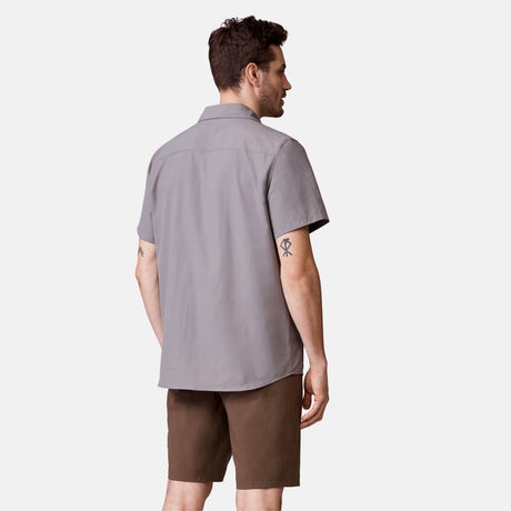 A man with short dark hair and a small upper right arm tattoo wears the Clearance Men's Expedition Nylon Rip-Stop Short Sleeve Shirt in light gray, paired with brown shorts, standing against a plain white background.