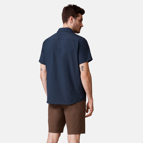 A man faces away, wearing the Clearance Men's Expedition Nylon Rip-Stop Short Sleeve Shirt in navy and brown shorts. His relaxed arms reveal small black tricep tattoos. The background is plain and light-colored.