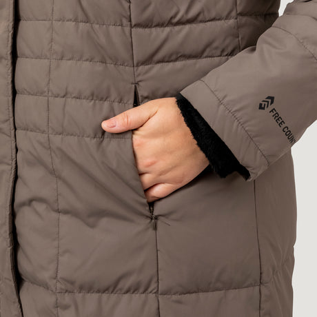 Woman wearing Free Country Women's Plus Size Full Length Splendor Down Jacket in brown with a full front zipper opening, lower pockets, calf length, a detachable hood with faux fur trim, and water resistant and wind resistant stretch performance fabric, light grey background. Best women’s jacket for winter weather.