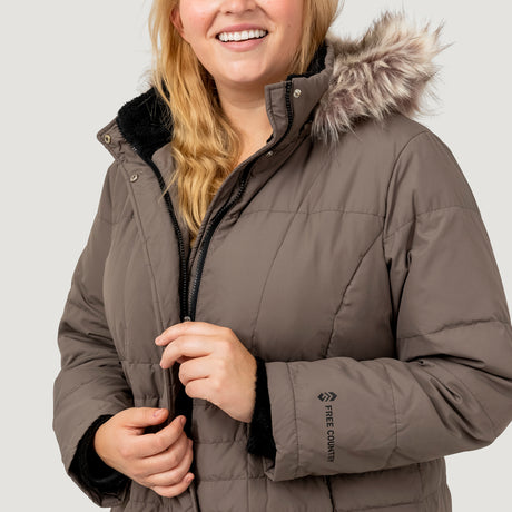 [Lola is 5'10.5" wearing a size 1X.] Woman wearing Free Country Women's Plus Size Full Length Splendor Down Jacket in brown with a full front zipper opening, lower pockets, calf length, a detachable hood with faux fur trim, and water resistant and wind resistant stretch performance fabric, light grey background. Best women’s jacket for winter weather.