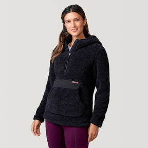 A woman with long brown hair wears a black Women's Sierra Butter Pile® Pullover Jacket by Clearance, featuring a front pocket and half-zip, paired with maroon pants as she smiles against a plain light gray background.