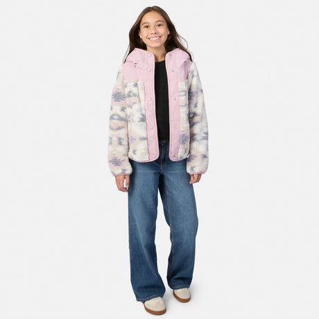 A young girl stands against a light gray studio background wearing a fuzzy hooded jacket with a pink hood and collar, a pastel tie-dye pattern on the sleeves and body, a black top, wide-leg blue jeans, and white slip-on shoes.